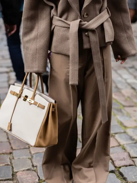 A woman in a camel coat carrying a white and gold Hermes Birkin on a cobbled street